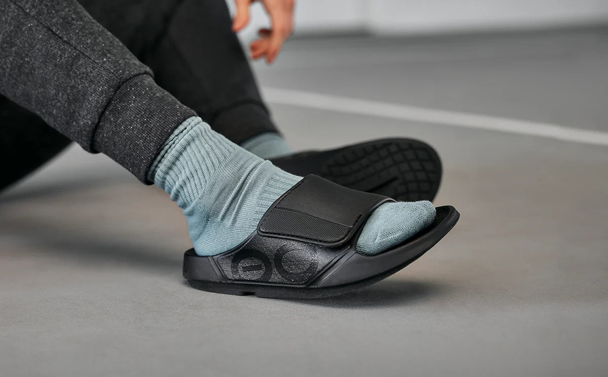 Easing a foot into an OOFOS Sport Flex while seated, showing how far the strap opens