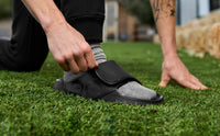 Pulling the adjustable strap of an OOFOS Sport Flex tight, crouched outdoors on grass