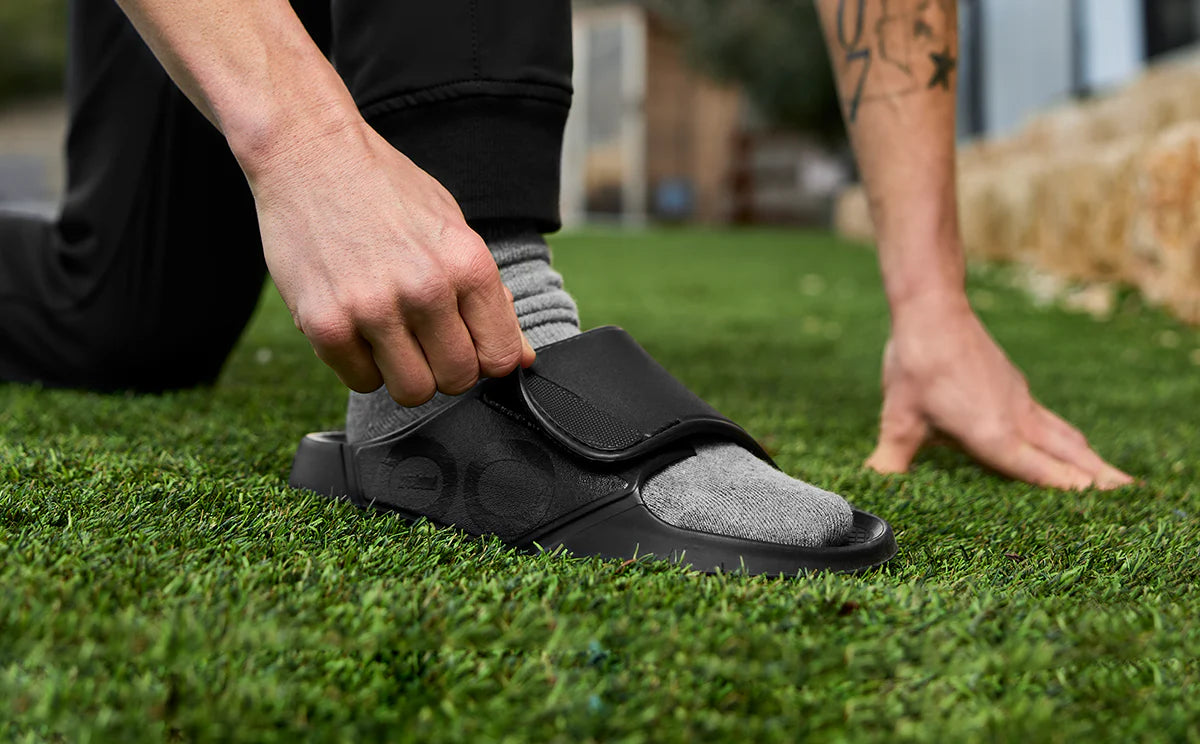 Pulling the adjustable strap of an OOFOS Sport Flex tight, crouched outdoors on grass
