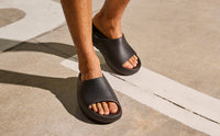 Black OOFOS OOahh Slide worn barefoot outdoors, showing how the sandal sits on the foot
