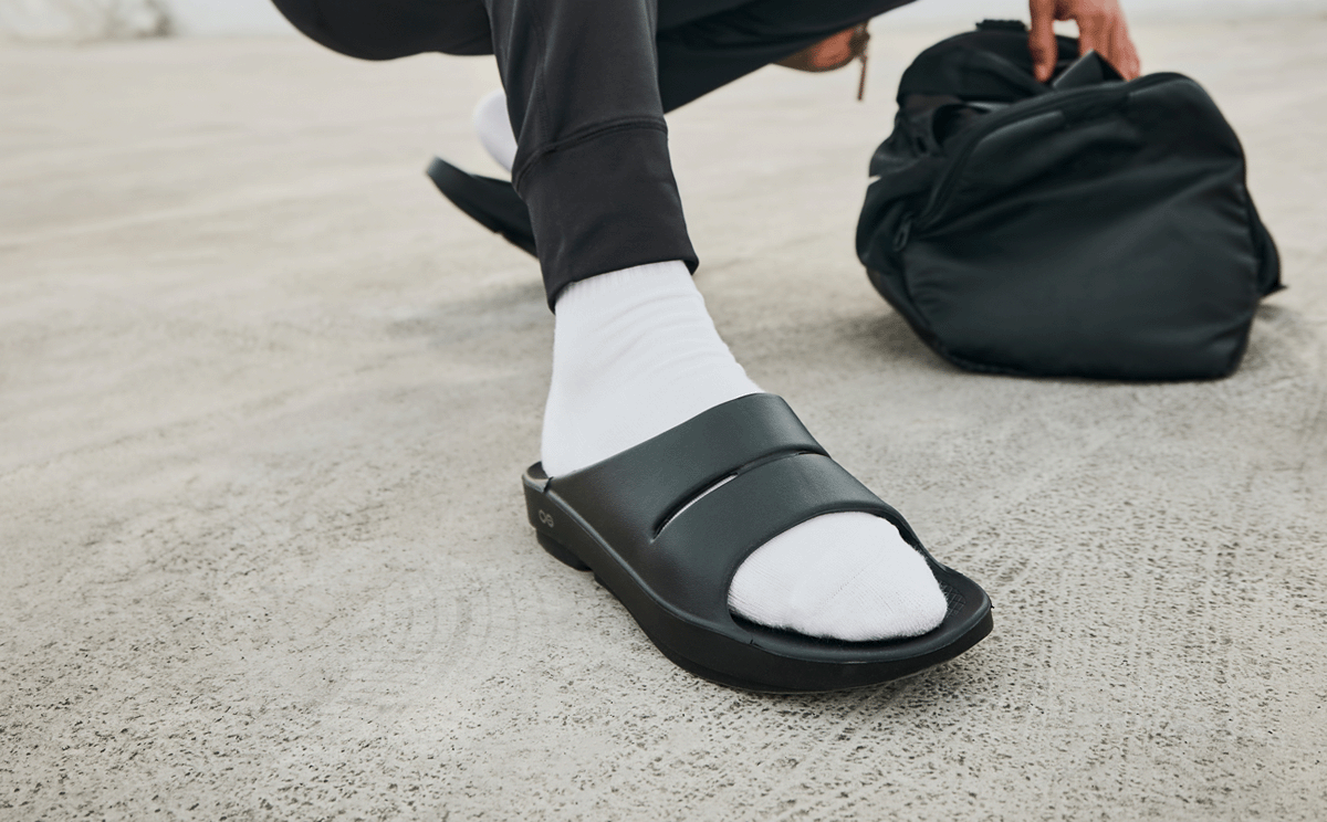 Black OOFOS OOahh Slide worn after a workout, man crouched on concrete beside a gym bag