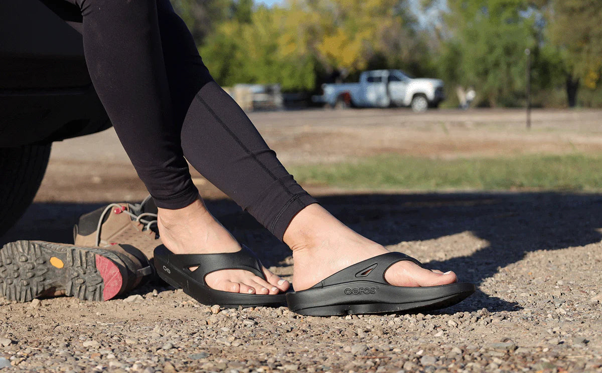 OOFOS OOriginal sandals worn after a run, crouched on gravel with a running shoe kicked off