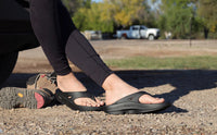 OOFOS OOriginal sandals worn after a run, crouched on gravel with a running shoe kicked off
